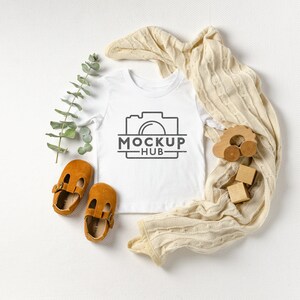 May include: White baby onesie with a black camera graphic and the text "MOCKUP - HUB". The onesie is laid flat on a white surface with a beige knitted blanket, a sprig of eucalyptus, and wooden toys.