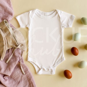 May include: A white baby bodysuit with short sleeves and a round neckline, next to a beige bonnet with bunny ears and a pink cloth. Several dyed eggs in shades of brown, green, and blue are scattered around the bodysuit.