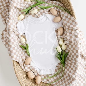 May include: A white baby onesie on a brown and white checked fabric in a wicker basket with white tulips and wooden eggs.