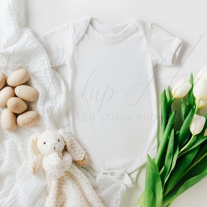 May include: A white baby onesie with a white crocheted blanket, wooden eggs, and a white plush bunny toy. A bouquet of white tulips with green stems is in the bottom right corner.