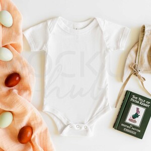 May include: A white baby onesie, a brown linen bonnet, a peach-coloured muslin swaddle, and a green book titled "The Tale of Peter Rabbit" by Beatrix Potter.  There are also several brown and green speckled eggs scattered around.