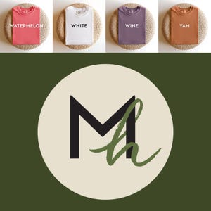 May include: Four folded t-shirts in watermelon, white, wine, and yam colors are displayed on woven mats. The shirts are arranged above a circular logo with a black "M" and a green "h" design on a cream background, set against a dark green backdrop.