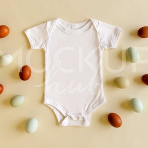 May include: A white baby onesie with brown, green and blue speckled eggs scattered around it on a light yellow background.