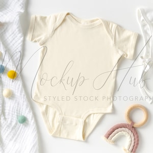 May include: A cream-coloured baby onesie with short sleeves, a white crocheted baby bonnet, a rainbow-coloured yarn and wooden ring rattle, and a white blanket with colourful pom-poms.