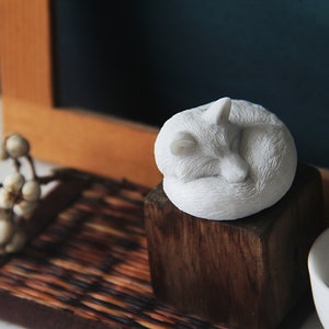 May include: White ceramic sculpture of a fox curled up sleeping on a wooden block. The fox is on a brown woven mat with a white flower sprig.
