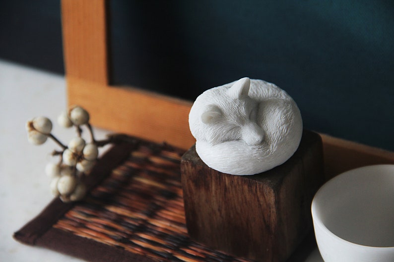 May include: White ceramic sculpture of a fox curled up sleeping on a wooden block. The fox is on a brown woven mat with a white flower sprig.