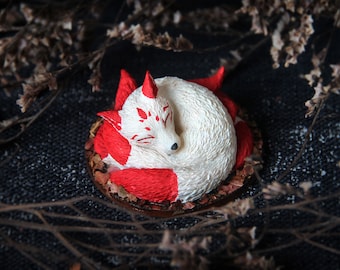 May include: A white and red ceramic fox figurine curled up sleeping. The fox has red markings on its tail and face. The figurine is sitting on a bed of brown leaves.