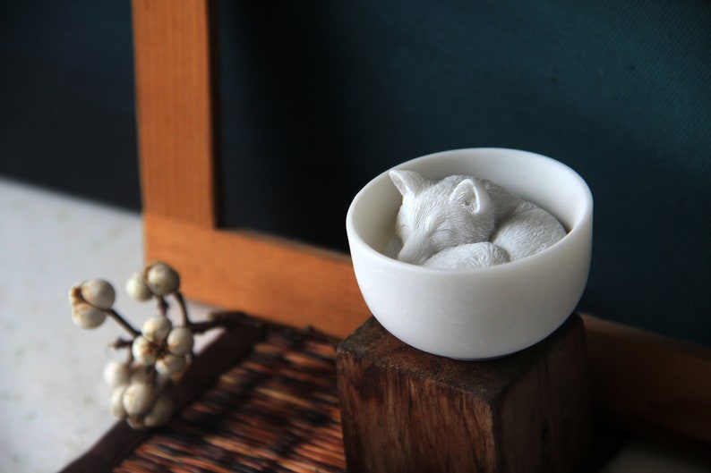 May include: A white ceramic bowl with a sleeping white fox figurine inside. The bowl is sitting on a wooden block.