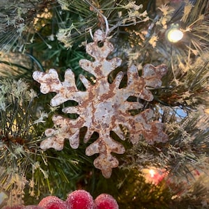May include: A rustic metal snowflake Christmas ornament with a weathered, brown and silver finish. The snowflake is hanging on a green Christmas tree with red berries and lights, creating a festive holiday scene.