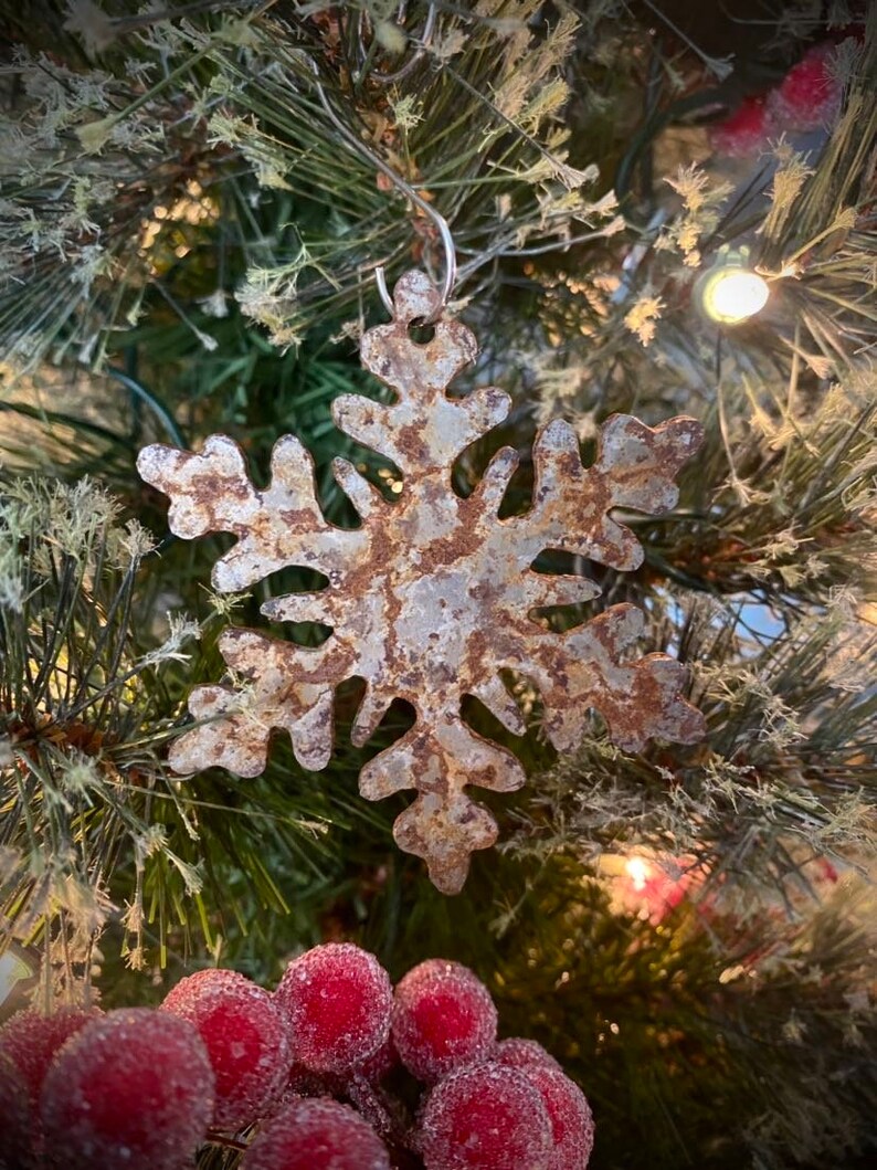 May include: A rustic metal snowflake Christmas ornament with a weathered, brown and silver finish. The snowflake is hanging on a green Christmas tree with red berries and lights, creating a festive holiday scene.