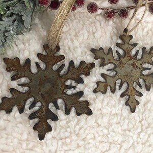 May include: Two rustic metal snowflake ornaments with a distressed finish. The snowflakes are in varying sizes and have a brown and gray color. Each ornament has a loop for hanging with a gold ribbon or twine. Decorative winter holiday decor.