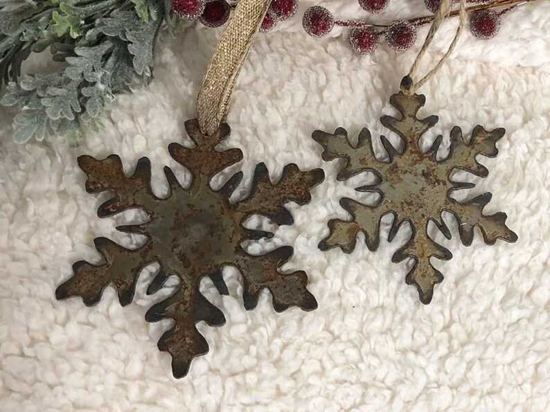 May include: Two rustic metal snowflake ornaments with a distressed finish. The snowflakes are in varying sizes and have a brown and gray color. Each ornament has a loop for hanging with a gold ribbon or twine. Decorative winter holiday decor.