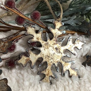 May include: Close-up of a rustic metal snowflake ornament with a distressed, aged appearance. The snowflake is off-white with blue-gray edges, and is hanging by a twine loop. Decorative red berries and greenery are in the background.