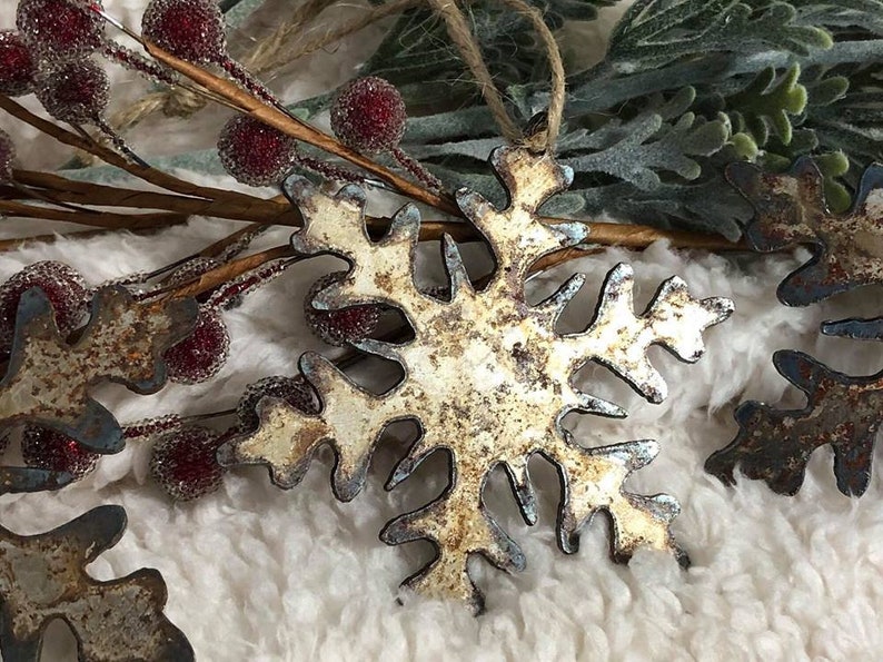 May include: Close-up of a rustic metal snowflake ornament with a distressed, aged appearance. The snowflake is off-white with blue-gray edges, and is hanging by a twine loop. Decorative red berries and greenery are in the background.