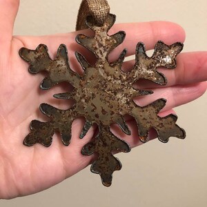 May include: A rustic metal snowflake ornament with a distressed, aged appearance. The snowflake is brown with darker edges and a textured surface. It has a loop of gold ribbon for hanging. A decorative winter holiday item.