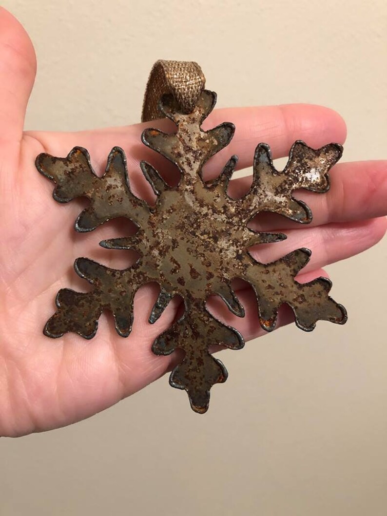 May include: A rustic metal snowflake ornament with a distressed, aged appearance. The snowflake is brown with darker edges and a textured surface. It has a loop of gold ribbon for hanging. A decorative winter holiday item.