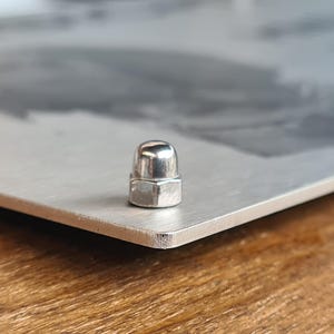 May include: A silver metal cap with a rounded top and a hexagonal base. The cap is attached to a metal surface.