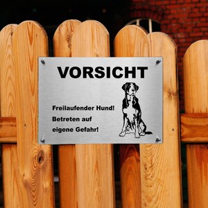 May include: A silver metal sign with black text that reads "Vorsicht Freilaufender Hund! Betreten auf eigene Gefahr!"  The sign also features a black and white illustration of a dog.