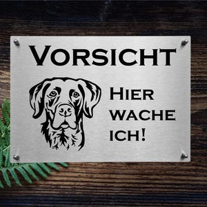 Könnte beinhalten: Ein weißes Metallschild mit schwarzem Text und einer schwarzen Silhouette eines Hundekopfes. Das Schild lautet "Vorsicht Hier Wache Ich!"