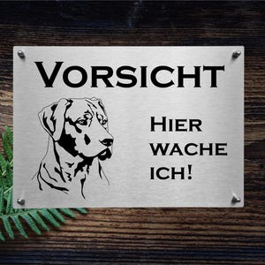 May include: A silver metal sign with black text and a black silhouette of a dog. The sign reads "Vorsicht Hier Wache Ich!" which translates to "Caution, I'm guarding here!"