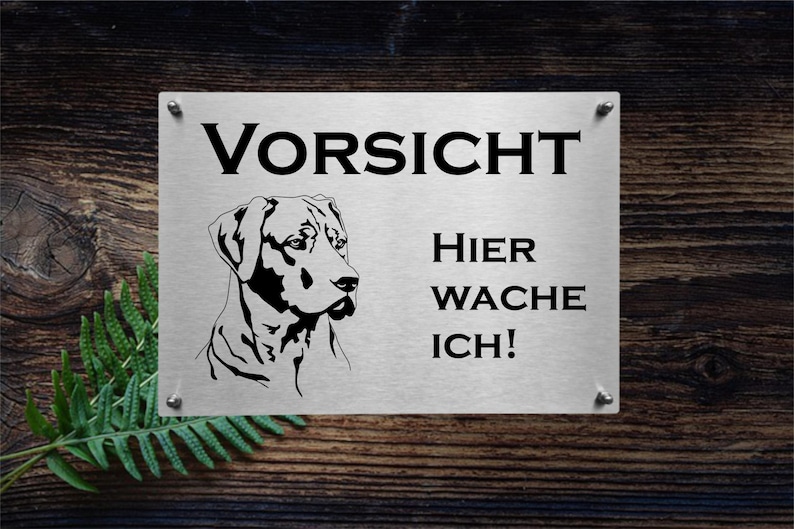 May include: A silver metal sign with black text and a black silhouette of a dog. The sign reads "Vorsicht Hier Wache Ich!" which translates to "Caution, I'm guarding here!"