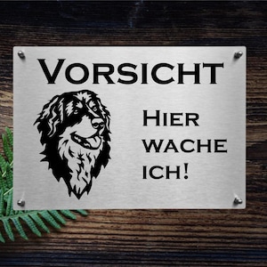 May include: A silver metal sign with black text and a black silhouette of a dog. The sign reads "Vorsicht Hier Wache Ich!" which translates to "Caution, I'm guarding here!"