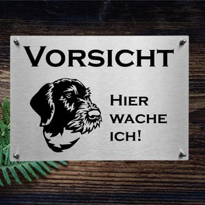 Könnte beinhalten: Ein weißes Metallschild mit schwarzem Text und einer schwarzen Silhouette eines Hundekopfes. Das Schild lautet "Vorsicht Hier Wache Ich!"