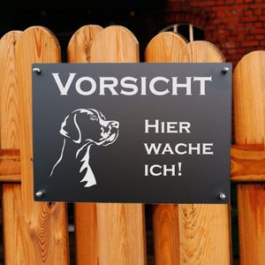 May include: A black rectangular sign with white text and a dog silhouette is mounted on a wooden fence. The sign reads "VORSICHT" at the top, with "Hier Wache Ich!" below, indicating a warning. The fence is made of vertical wooden planks.