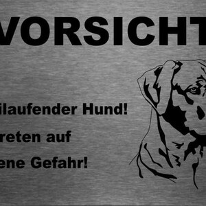 May include: A silver metal sign with black text that reads "Vorsicht Freilaufender Hund! Betreten auf eigene Gefahr!"  The sign also features a black silhouette of a dog's head.