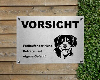 Könnte beinhalten: Ein silbernes Metallschild mit schwarzem Text, der "Vorsicht Freilaufender Hund! Betreten auf eigene Gefahr!" lautet. Das Schild zeigt außerdem eine schwarze Silhouette eines Hundekopfes.