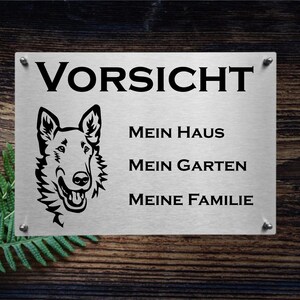 May include: A silver metal sign with black text and a black silhouette of a dog's head. The sign reads "Vorsicht", "Mein Haus", "Mein Garten", and "Meine Familie".