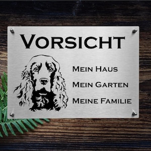 Könnte beinhalten: Rechteckiges Metallschild mit dem Wort "VORSICHT", einer Hundeillustration und Text. Das Schild ist auf einem Holzgrund befestigt. Der Text enthält "MEIN HAUS", "MEIN GARTEN" und "MEINE FAMILIE".