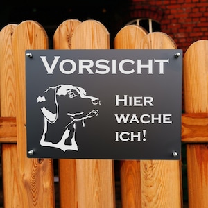 Könnte beinhalten: Ein schwarzes rechteckiges Schild mit weißem Text und einer Hunde-Silhouette, montiert an einem Holzzaun. Das Schild lautet "VORSICHT HIER WACHE ICH!" in weißen Großbuchstaben. Die Hunde-Silhouette ist ein Seitenprofil. Der Zaun besteht aus vertikalen Holzplanken.