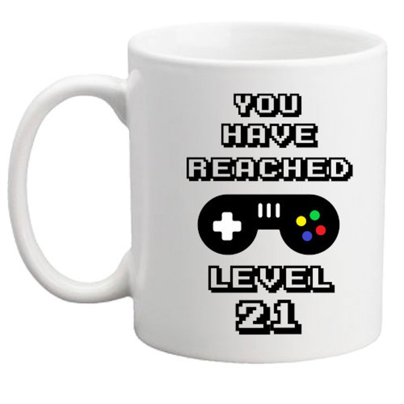 Może przedstawiać: Biały kubek ceramiczny z czarnym tekstem, kt&oacute;ry brzmi "You have reached level 21". Na kubku nadrukowano czarno-biały, pikselowany kontroler do gier wideo z zielonymi, czerwonymi i niebieskimi przyciskami.