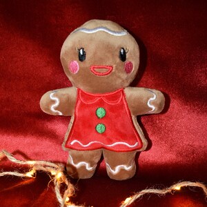 May include: A plush gingerbread person toy with a red dress and green buttons. The gingerbread person has white icing details, pink cheeks, and a smiling red mouth. The toy is set against a red velvet background, with a string of lights in the foreground.