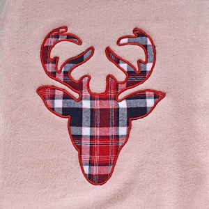 May include: A plush, light pink fabric with a deer head applique. The deer head is outlined in red and filled with a red, black, and white plaid pattern. The antlers are detailed and the overall design is centered.