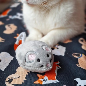 以下が含まれることがあります： 緑色の目をした三毛猫と灰色のネズミのおもちゃ。 猫は白、オレンジ、黒の毛皮をしています。 ネズミのおもちゃは灰色で、ピンクの耳と白い紐の尾が付いています。 猫は模様のある布の上にいます。