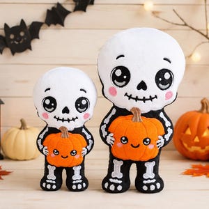 May include: Two plush skeleton dolls with white skulls, large black eyes, and pink cheeks, each holding a small orange pumpkin with a smiling face. The skeletons have black bodies with white bone details on the arms, legs, and torso, crafted in a soft, stuffed fabric style.