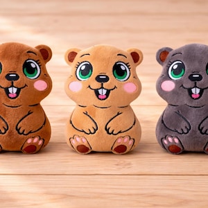 Peut inclure: Trois peluches de marmottes en marron, beige et gris. Chacune a de grands yeux verts, des joues roses et une bouche souriante. Elles ont de petites oreilles, des pattes et sont assises. Les marmottes sont sur une surface en bois.