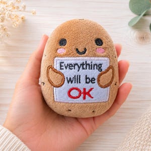 Peut inclure: Une peluche marron en forme de pomme de terre avec des traits brodés, incluant des yeux noirs et des joues roses. Elle tient un panneau rectangulaire blanc avec le texte noir brodé Everything will be et le grand texte rouge OK.