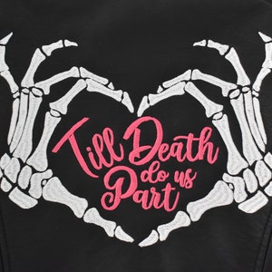 Może przedstawiać: Czarna kurtka z sercem utworzonym z białych szkieletowych dłoni. Różowy haft głosi "Till Death do us Part". Kurtka ma gładką, skóropodobną fakturę. Motyw gotycki.