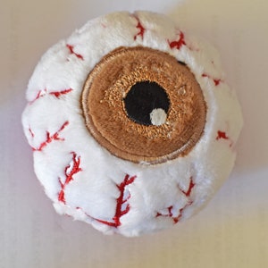 Może przedstawiać: Pluszowa zabawka w kształcie oka, o okrągłym kształcie, na białym tle. Posiada brązową tęczówkę, czarną źrenicę i czerwone żyły. Zabawka wykonana jest z miękkiego, pluszowego materiału.
