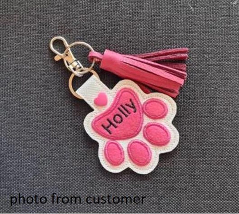 May include: A personalized pet paw print keychain with the name "Holly" embroidered in black on a pink paw print design. The keychain includes a pink tassel and a silver-colored clasp.