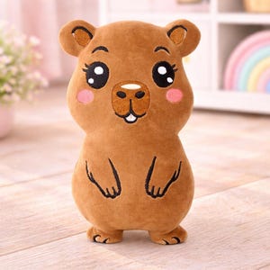 Peut inclure: Une peluche marron représentant un capybara, avec de grands yeux expressifs, des joues roses et une bouche souriante. Le jouet a de petites oreilles et des détails brodés en noir. Il se tient debout sur une surface claire.