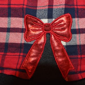 May include: Close-up of red, white, and navy blue plaid fabric with a large, shiny red bow applique. The bow is centered, adding a festive touch to the plaid pattern.