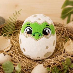 May include: A plush toy shaped like a cracked egg with a green dinosaur face peeking out. The egg is off-white with light brown spots, while the dinosaur features large black eyes with white highlights and a green snout with embroidered details.