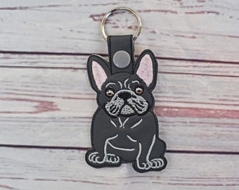 May include: Black French Bulldog keychain with a silver keyring. The dog has white embroidered details and pink inner ears. The keychain is made of a leather-like material and features a snap closure.