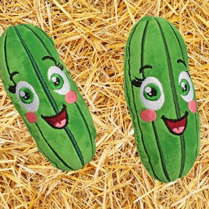 May include: Two plush green pickle toys with smiling faces, rosy cheeks, and large eyes. The pickles have vertical lines and are set against a background of straw. These whimsical toys are designed for children.