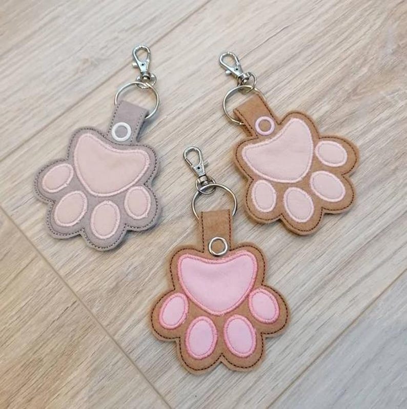 May include: Three paw-shaped keychains in shades of gray, tan, and brown, each with a pink paw pad design. The keychains have silver-colored metal hardware and a loop for attaching keys.