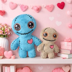 May include: Two plush voodoo dolls, one blue and one tan, with stitched features and heart-shaped accents. The dolls have large, expressive eyes and stitched smiles. They are displayed on a white shelf with pink heart decorations in the background.